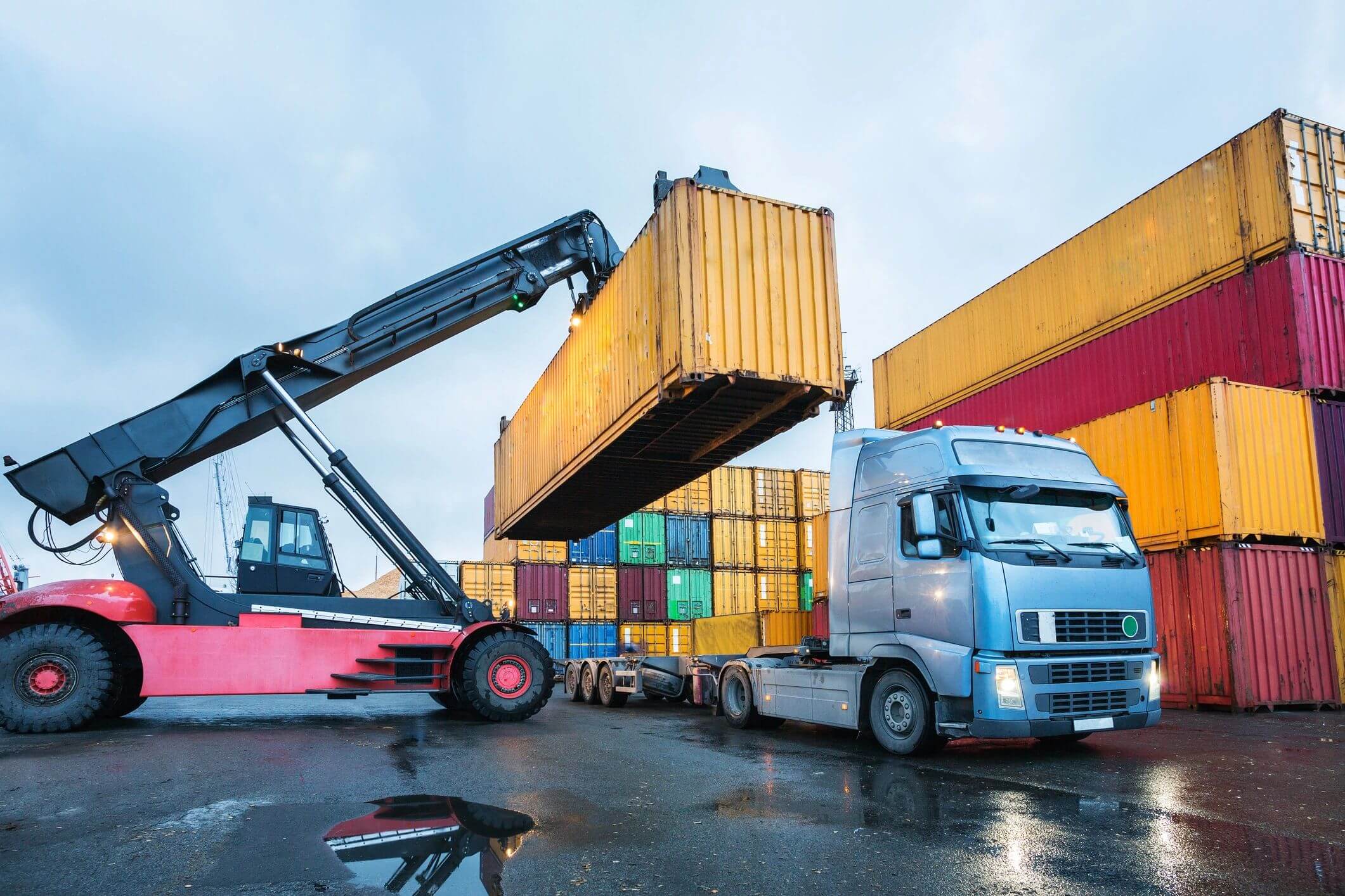 Crane loading a shipping container onto a truck in a busy port yard.