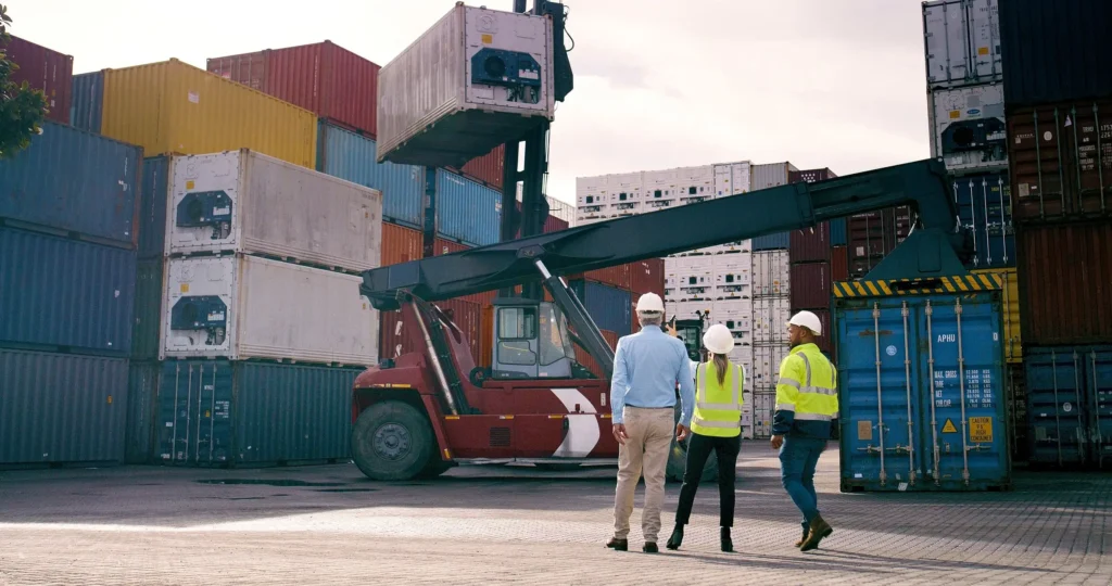 Workers inspecting a large container handler at a shipping yard.